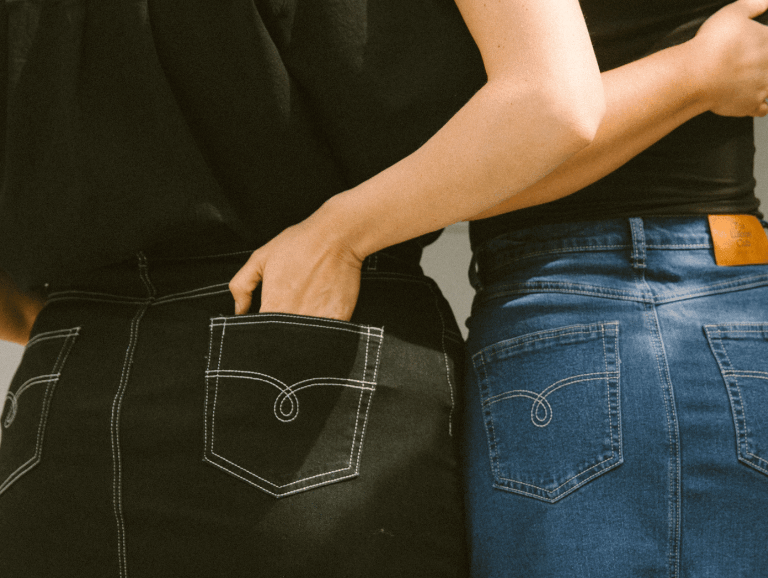 Two women standing next to each other with their hands in each others jean pockets. It's a close up of two pairs of jeans, blue and black.