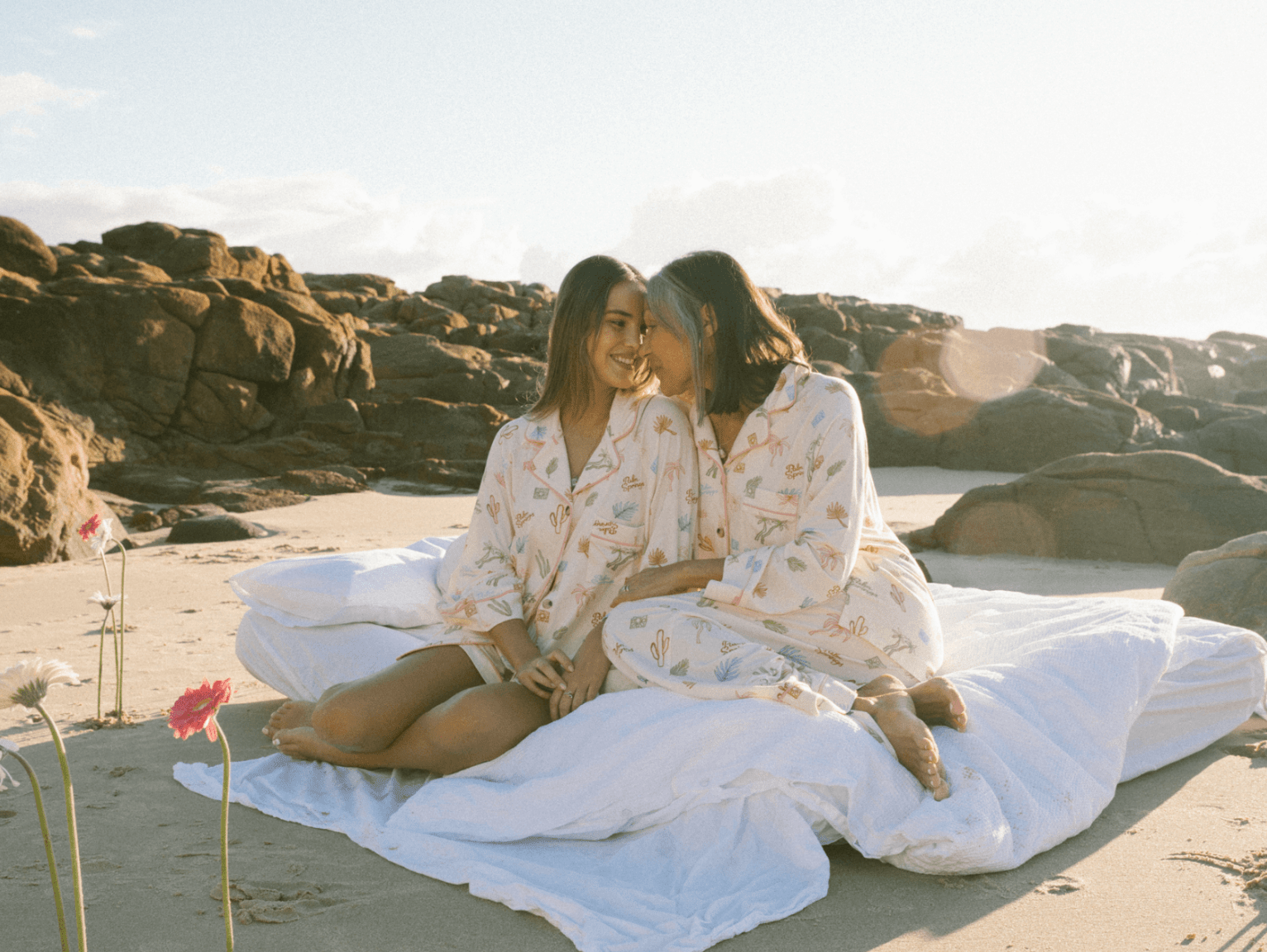 A mother and daughter wearing cosy pyjamas sitting on a bed on a sunny beach in the morning, surrounded by flowers