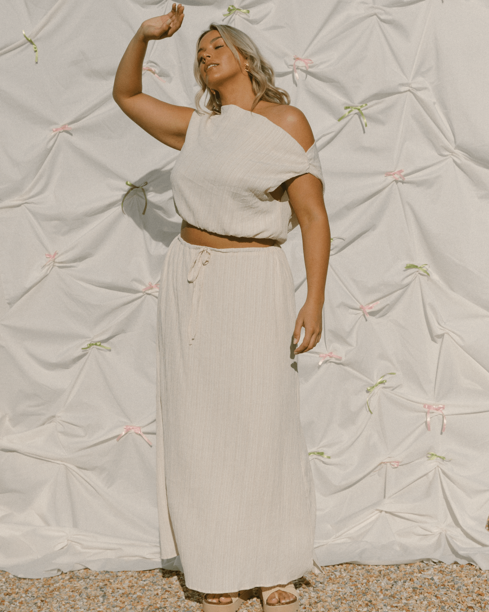 Woman in a beige outfit standing against a textured white wall with decorative elements.
