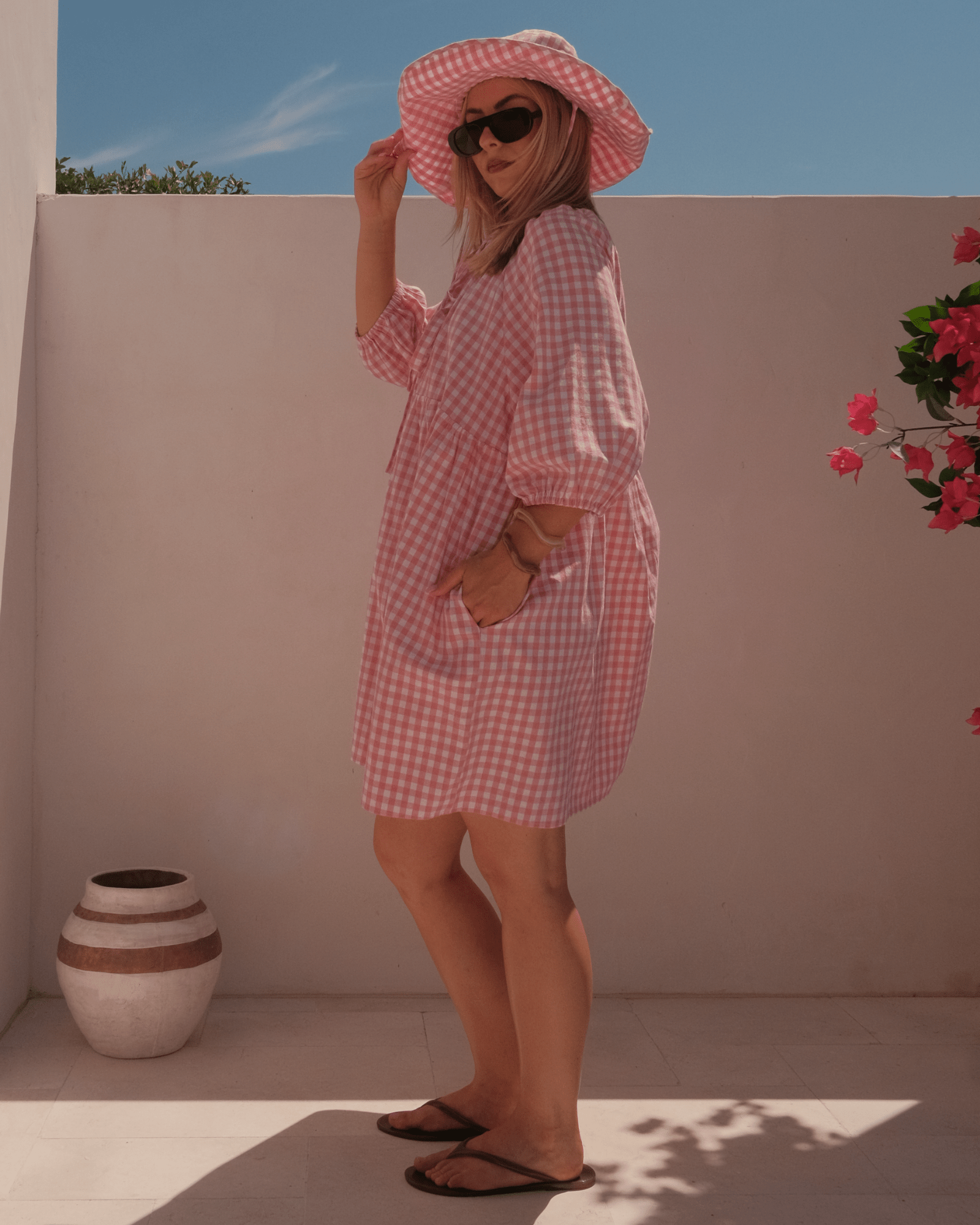 Woman in a pink checkered dress and hat standing against a white wall with a vase and flowers.