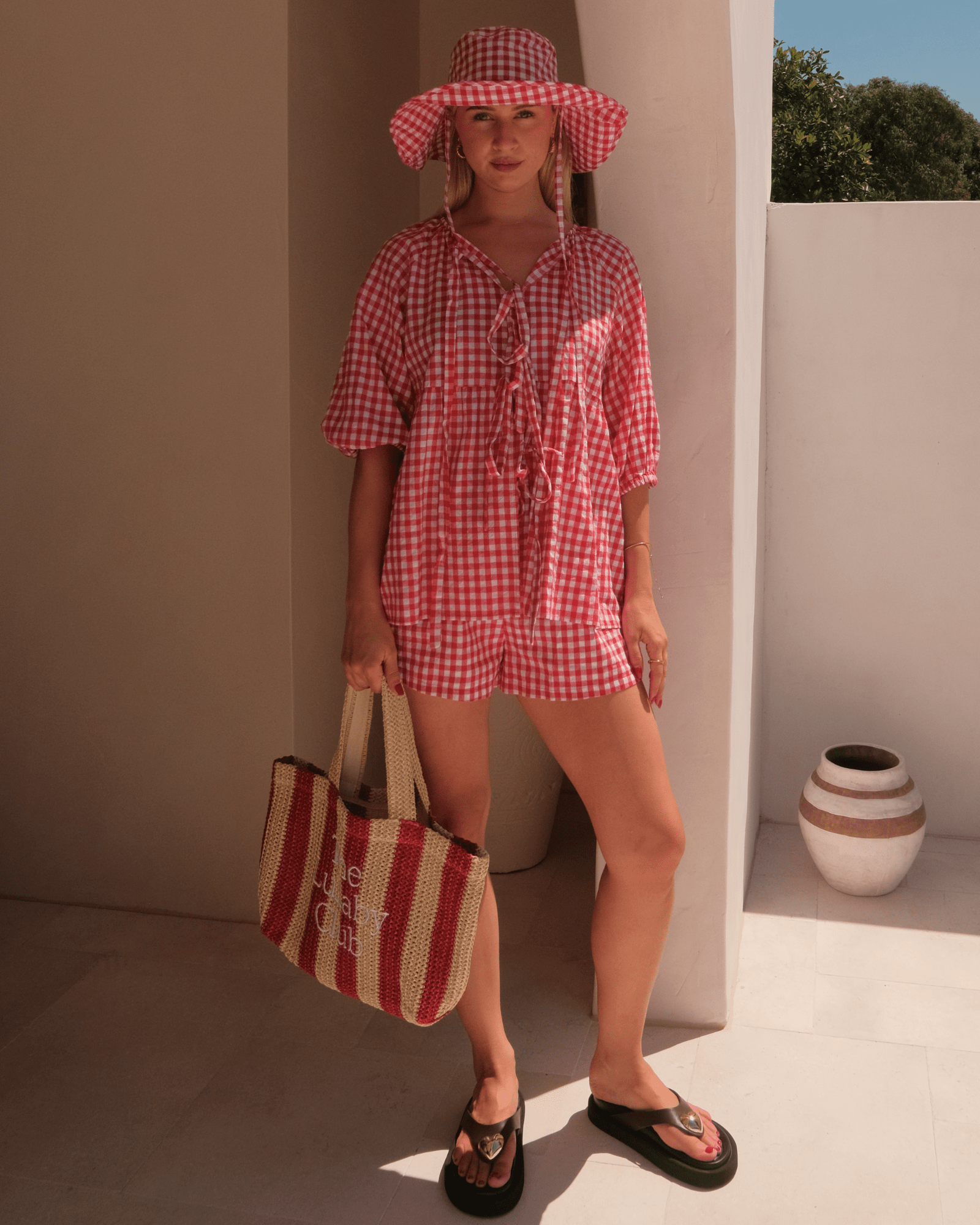 Woman in a red and white checkered outfit holding a striped bag and a matching hat.
