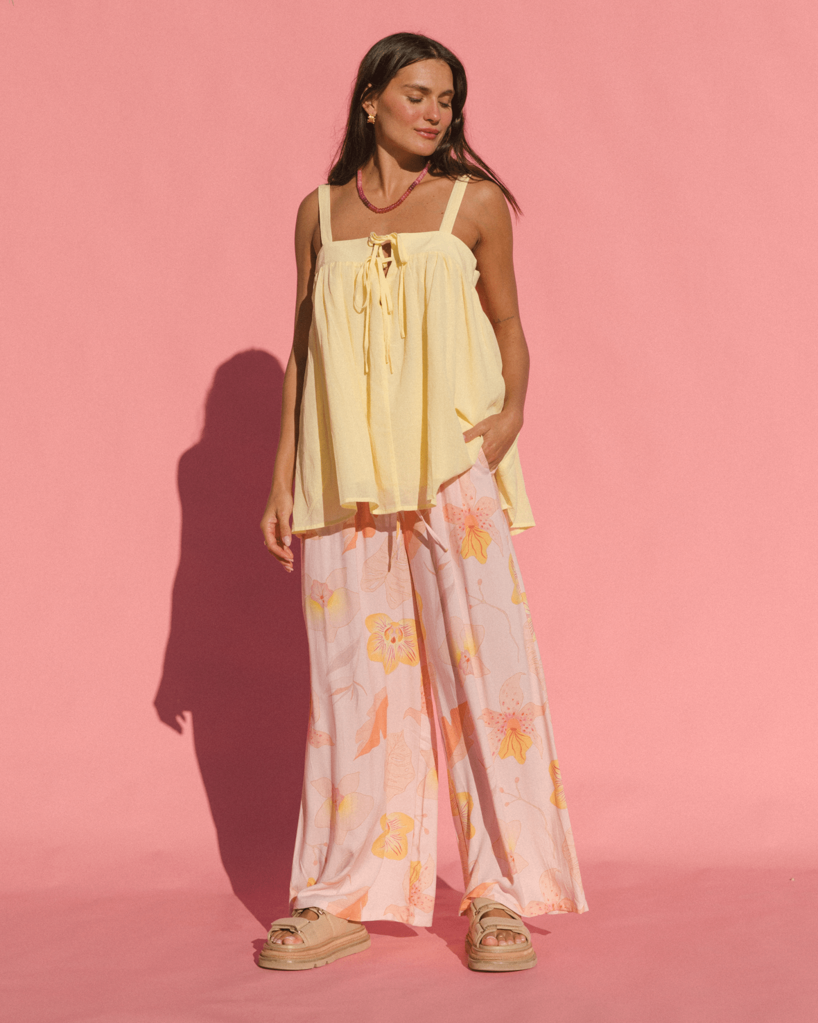 Woman wearing a flowy yellow cotton top and light pink floral pants against a pink background
