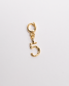 A gold-colored charm in the shape of the number 5 with a secure clasp, designed for attachment to jewelry such as necklaces and bracelets.