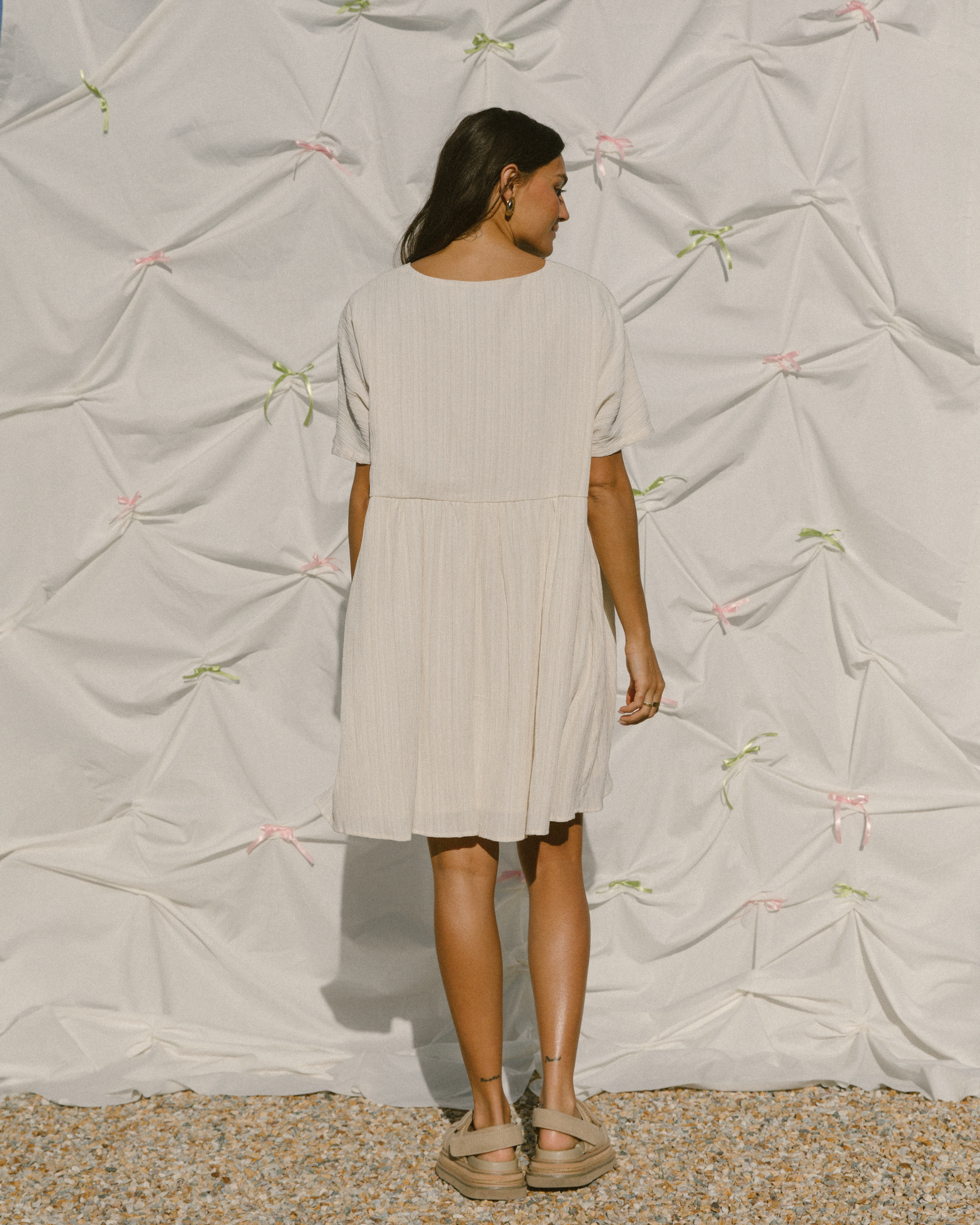 Woman wearing a light coloured smock style mini dress in a plisse fabric. 