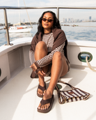 A woman wearing sunglasses, a brown vest, and a brown-and-white striped long-sleeve top sits on the deck of a white boat, with a matching striped bag beside her.