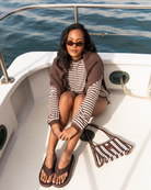 A woman wearing sunglasses, a brown and white striped long-sleeve top, and brown flip-flops sits on the white deck of a boat with blue water in the background. A matching brown and white striped handbag rests beside her.