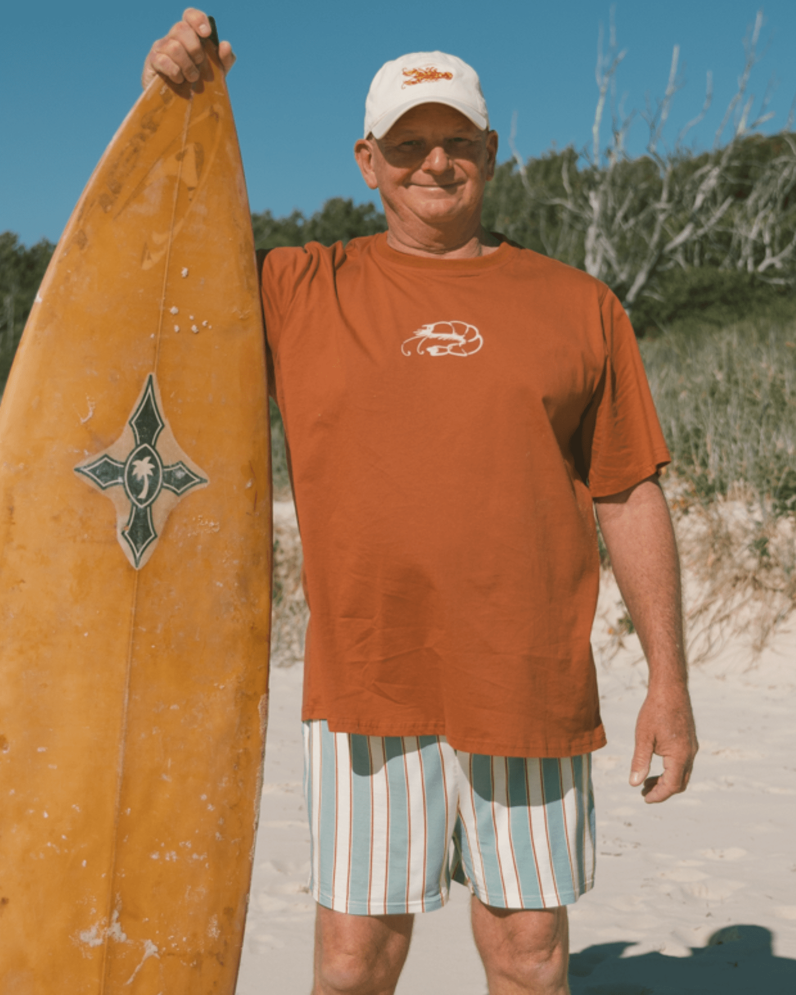 Man holding a yellow surfboard on a beach in matching rust pj set. 