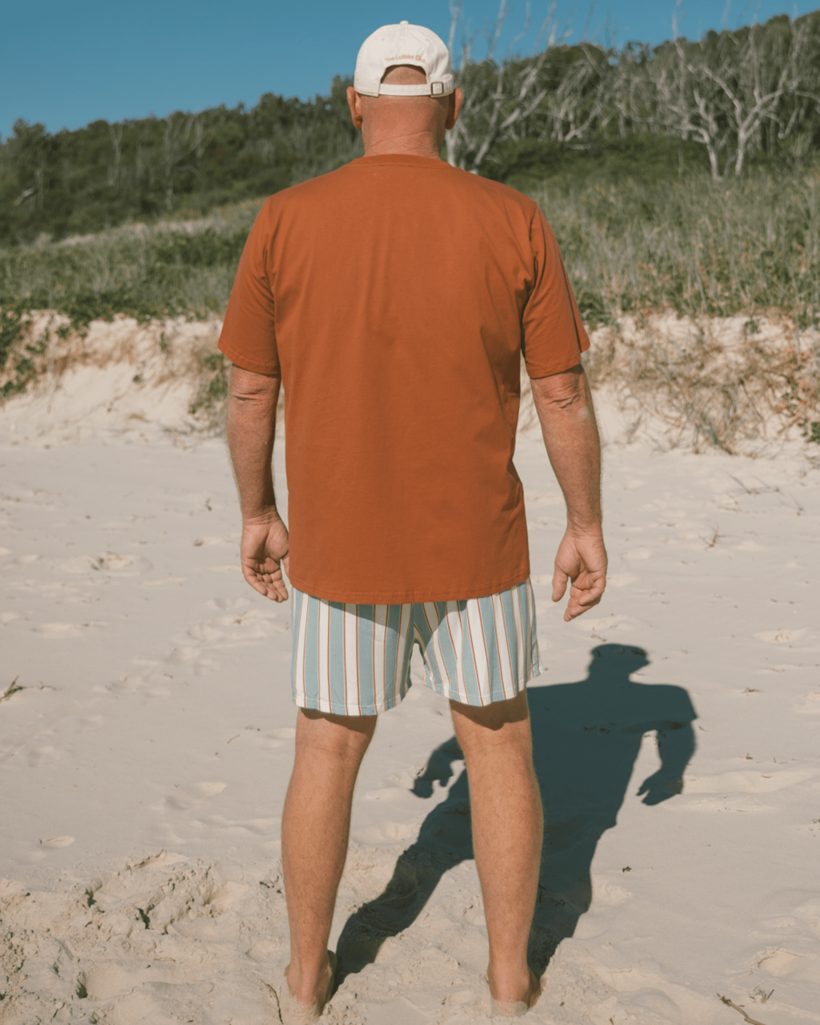 Man walking on a sandy beach wearing an orange shirt and striped shorts.