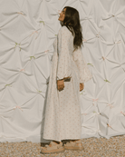 Woman in a long, light-colored dress standing against a textured white wall with decorative elements.
