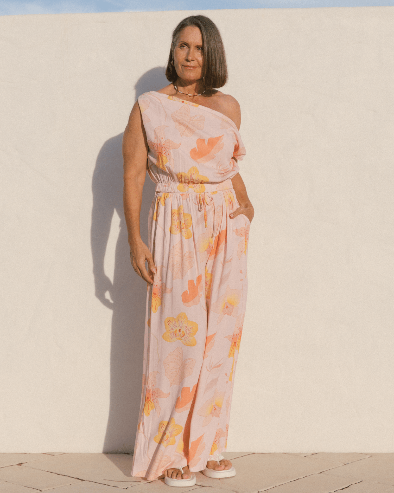 Woman wearing off the shoulder crop top and matching wide legged pants in a light pink floral design. 