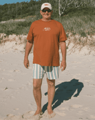 Man standing on a sandy beach wearing an orange t-shirt and striped shorts.