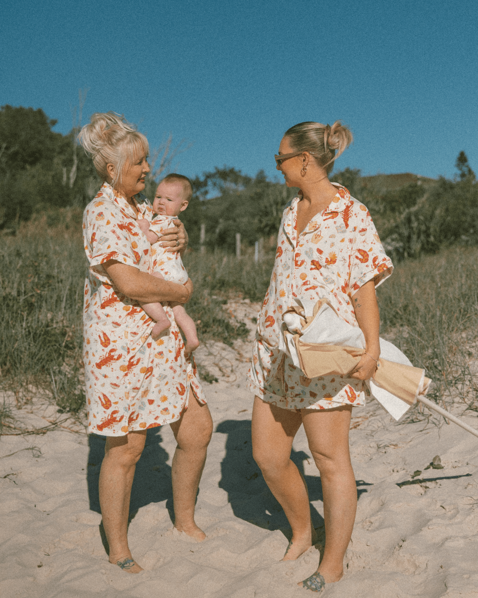 Two women in matching christmas outfits standing on a sandy beach with a baby.