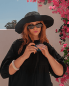 Person wearing a black hat and sunglasses, holding a colorful object, with pink flowers in the background.