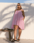 Woman in a pink dress standing against a white wall with a wooden bench and black and white bag.