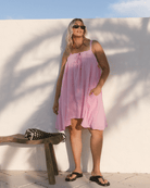 Woman in a pink dress standing against a white wall with a wooden bench and black and white bag.